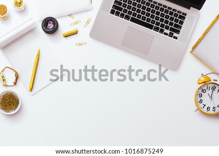 Desktop flatlay scene, laptop, notepad, pens along with other gold stationery items, on a plain white desk background. Negative space below.