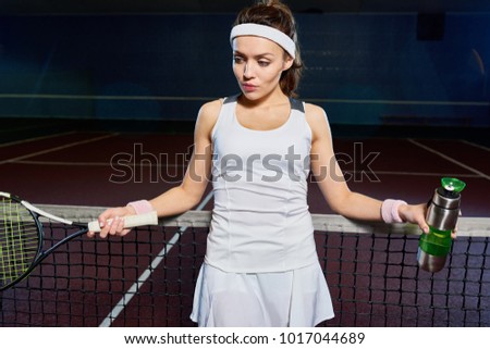 Portrait of female tennis player leaning on net and holding water bottle taking break from practice in indoor court, copy space