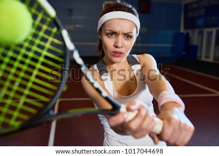 Portrait of forceful woman playing tennis in indoor court, frowning with strain while hitting ball with racket , shot with flash