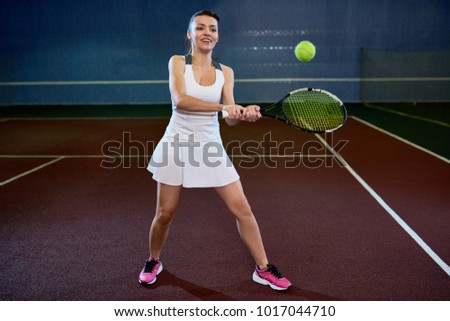 Full length portrait of joyful young woman playing tennis in indoor court, ready to hit flying ball and smiling happily, copy space