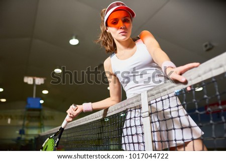 Low angle portrait of pretty young woman posing with net at indoor tennis court, looking at camera, copy space