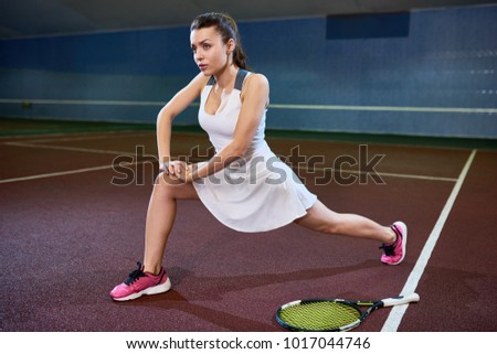 Full length portrait of female tennis player warming up and stretching before practice in indoor court, copy space