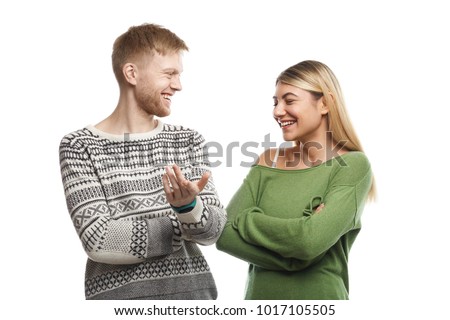 Picture of charismatic guy with stubble smiling happily while telling funny story to attractive young female with fair hair who laughing at his jokes. Cute couple talking against white wall background