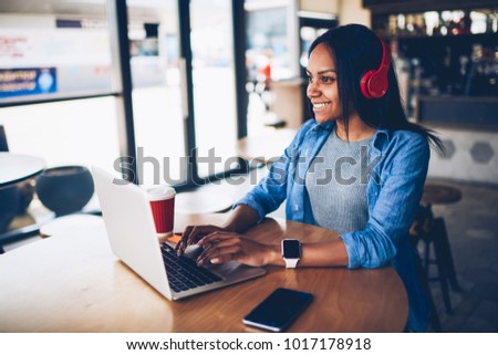Cheerful female black model resting in coffee shop listening favorite music in headphones and chatting in networks.Beautiful dark skinned freelancer enjoying remote job typing publication on laptop