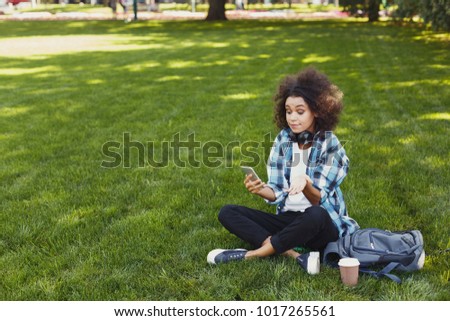 Embarrassed african-american girl waiting for call. Young woman looking hopefully on smartphone, sitting with headphones on grass at city park. Technology and leisure concept