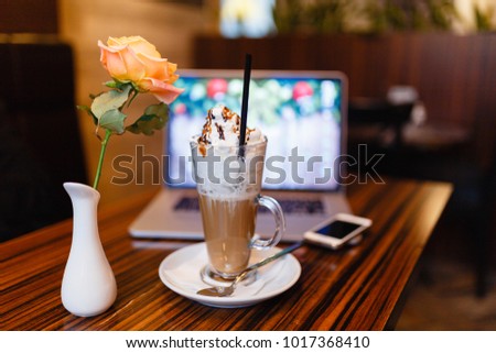 Glass Cup of coffee latte with cream and marshmallows and a pink rose in a vase on the background of laptop and smartphone in a cozy cafe. Iced Cafe Latte or Cappuccino with computer notebook