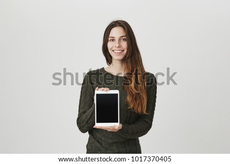 Shot of joyful beautiful dark-haired girl in green sweater standing against gray background with smile, demonstrating screen of tablet computer. Satisfied customer. Advertising concept