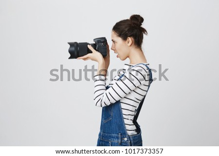 Profile of hipster girl holding photo camera in hands, taking photos. Brunette photographer with two hairbuns in denim overall concentrated on her work, posing against gray background