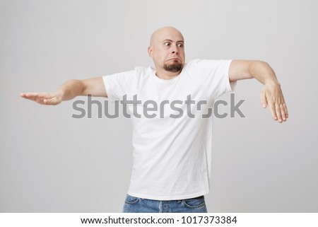Indoor portrait of funny bald bearded european guy who is doing wave dance while standing over gray background in white t-shirt and jeans. It is never too late to start learning how to breakdance