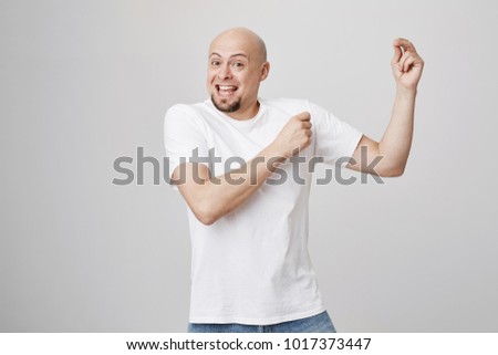 Indoor shot of happy positive bald man with beard, wearing white t-shirt and jeans being excited and overwhelmed with emotions while dancing cheerfully over gray background. Guy finally got divorce