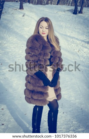 A young blonde girl is walking in the woods in winter. She is wearing a fur coat. Girl model posing among trees and snow