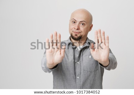 Body language. Disgusted serious angry bald man with beard in grey shirt posing against studio wall, keeping hands in stop gesture, trying to defend himself as if saying: Stay away from me Royalty-Free Stock Photo #1017384052