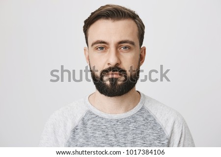 Calm interesting bearded guy in trendy shirt with normal face expression, looking at camera while standing against gray background. Man takes photo for passport at local studio
