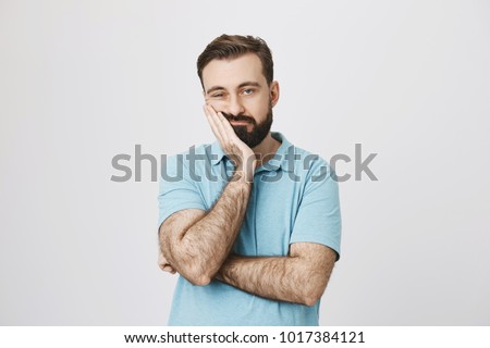 Very bored adult man with beard holding hand on cheek while support it with another crossed hand, looking tired and sick, over gray background. Father attends parents day at school Royalty-Free Stock Photo #1017384121