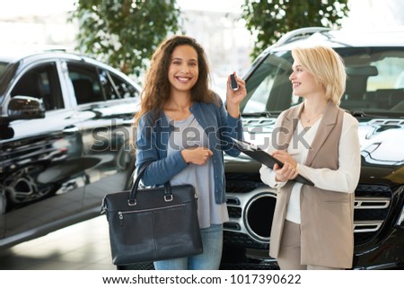 Attractive young customer showing key of new car to camera and posing for photography with toothy smile while blonde saleswoman standing next to her, interior of modern showroom on background