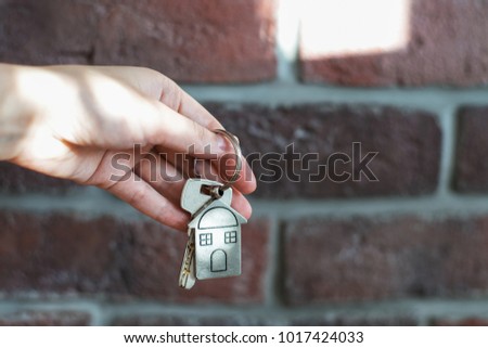 Female hand holds keys to the house on the brick wall background. Keys with a keychain in the form of a house. Home sales concept image. The keys to the property.