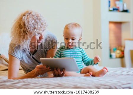 Little  infant baby boy with curly blond female nanny working with tablet while siting in bedroom at cozy home interior. Real people life and different generations concept.