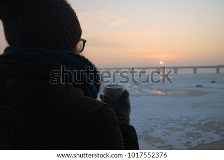 A girl holds a coffee paper cup in her hands and watches winter sunrise on the banks of the Dnieper River in Kyiv, Ukraine. Close-up photo.