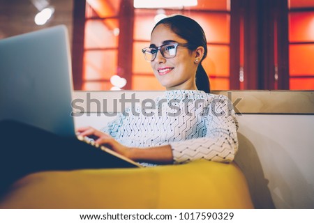 Smiling woman in eyewear watching video on laptop computer working on freelance in cozy coworking interior, female student rest during free time reading news blogging in social networks using netbook Royalty-Free Stock Photo #1017590329