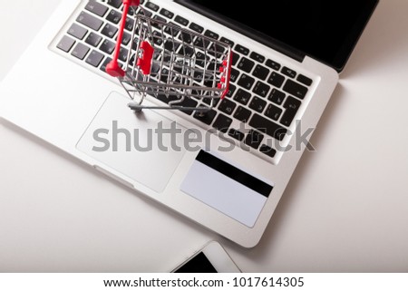 Online shopping. Bank card nearby a laptop and mini shopping cart on white background top view. 