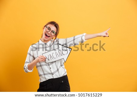 Portrait of funny woman in plaid shirt holding speech arrow pointer with word vacancy, isolated over yellow background Royalty-Free Stock Photo #1017615229