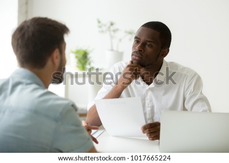 Serious attentive african hr manager listening to caucasian candidate at job interview, focused strict employer thinking having doubt about hiring decision while talking to male applicant in office Royalty-Free Stock Photo #1017665224