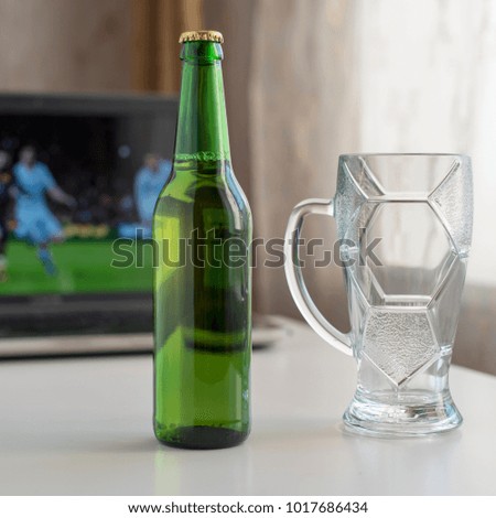 Bottle of light beer and empty beer glass mug on a white table. The background shows the game football on the laptop.
