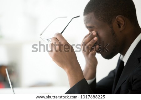 African tired businessman feels eyestrain taking off optical computer glasses, holding spectacles in hands massaging eyes, eyesight problem correction, fatigue, overwork and stress at work concept Royalty-Free Stock Photo #1017688225