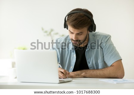 Focused man wearing headphones writing notes studying with laptop, serious student or office worker listening to audio business course at work, e-learning and online professional education concept Royalty-Free Stock Photo #1017688300