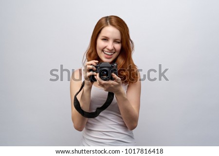 portrait of young attractive redhead girl photographer in a white t-shirt with camera in hand. Joyful expression, wide smile and winks at the camera. Close-up portrait on white background