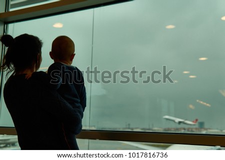 A mother and his little boy infant saying goodbye through an airport window in a melancholic mood; during an airplane take-off in a cloudy weather, while the father is departing to a new destination