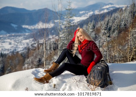 Winter mountain landscape photo of smiling beautiful blonde girl that sitting on the rock with the backpack.