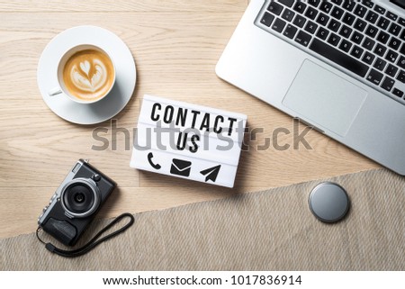 Contact us writing with emoticons in lightbox with camera, coffee mug and laptop lying on office desk as flat lay