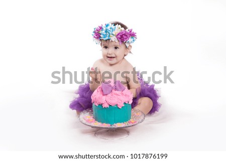 Cute happy baby girl with blue eyes is tasting the  cake and purple butter icing on her sticky fingers from her first birthday cake smash while sitting on a white background 
