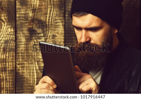 Bearded man, long beard. Brutal caucasian serious unshaven hipster holding tablet in black leather jacket on brown vintage wooden studio background