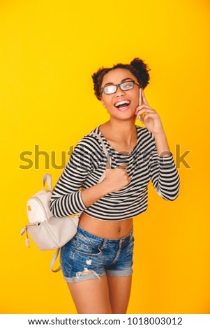 Young african woman isolated on yellow wall studio teen style with knapsack smartphone communication