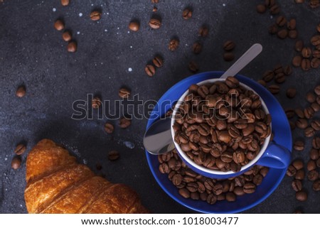 Cup with roasted coffee beans, spoon, croissant. View above.