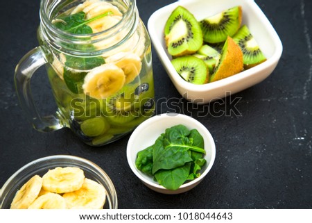 A jar of detox water with fresh fruit and ingredients on the table: chopped kiwi, sliced banana and spinach in bowls ,black stone background