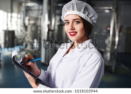 a pretty young girl scientist or worker in white uniform makes notes on paper against the background of modern factory equipment.