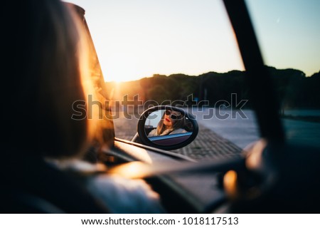 Reflection of young and fashionable woman in sideview mirror of small convertible cabriolet car, beautiful sun leaks and beams, fresh and sunny mood of summer vacation or holiday destination