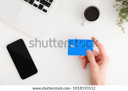 Man holding credit card over white office desk with smartphone and laptop. Top view.