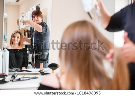 Close-up of professional hairdresser stylist drying her client's hair with a hairdryer after a haircut in beauty salon. Satisfied woman is looking at herself in a mirror.