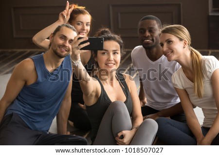 Sporty young fit smiling multi-ethnic people having fun taking group selfie in gym, diverse happy black and white friends making photo on smartphone together sitting in yoga studio at training break