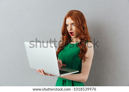 Photo of redhead young shocked confused lady in green dress standing over grey wall background using laptop computer looking aside.