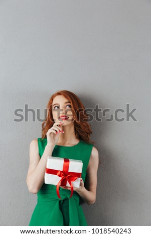 Image of thinking redhead young woman in green dress standing over grey wall background. Looking aside holding surprise gift box.