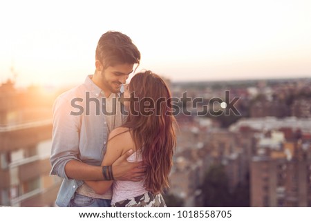 Couple in love standing and hugging on a building rooftop at sunset with cityscape in the background. Focus on the girl