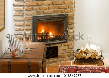Stylish interior of living room with fireplace, wine, glass, rug, tile floor and brick stone wall. Winter and Romance concept. Fire is burning and basket with firewood near.