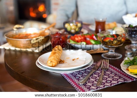 Delicious Turkish, Greek and Arabic Breakfast serving sliced black and green olive, black tea, tomato, cucumber, fried borek, arugula, bread and strawberry jam on table with fireplace background.