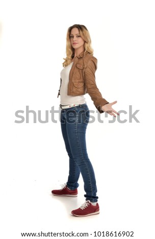 full length portrait of blonde lady wearing simple brown jacket and jeans, standing pose isolated on white studio background.