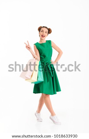 Full length portrait of a satisfied young girl dressed in green dress holding shopping bags isolated over white background